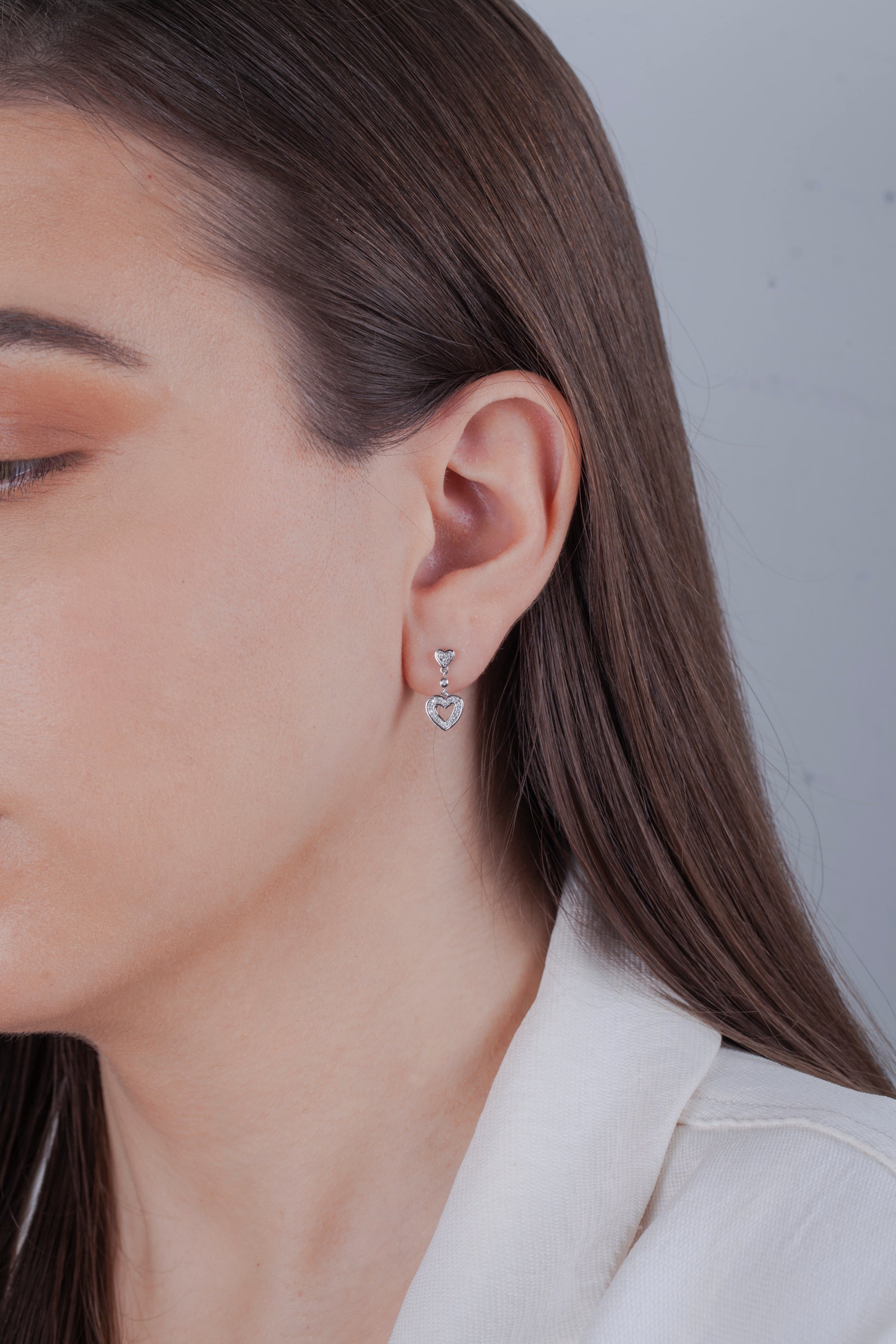 Close-up of a person wearing sterling silver dangle heart-shaped diamond earrings with a neutral background