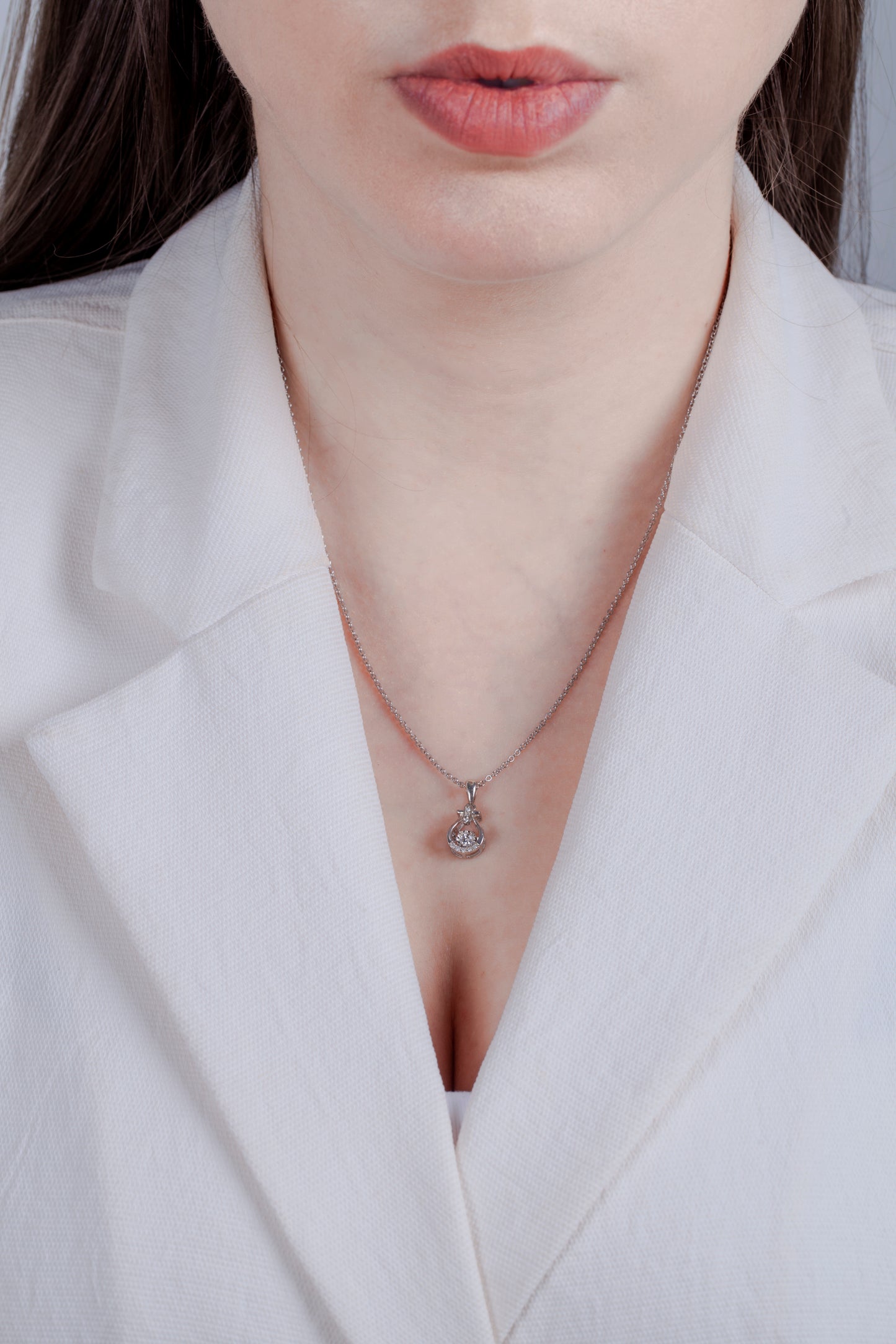 Model in white blazer wearing sterling silver pendant featuring a contoured bell-shaped frame with a structured crossbar at the top. The center holds a dancing solitaire diamond in a miracle setting, accented by three vertical diamonds on the bale and a curved row of diamonds along the bottom edge.