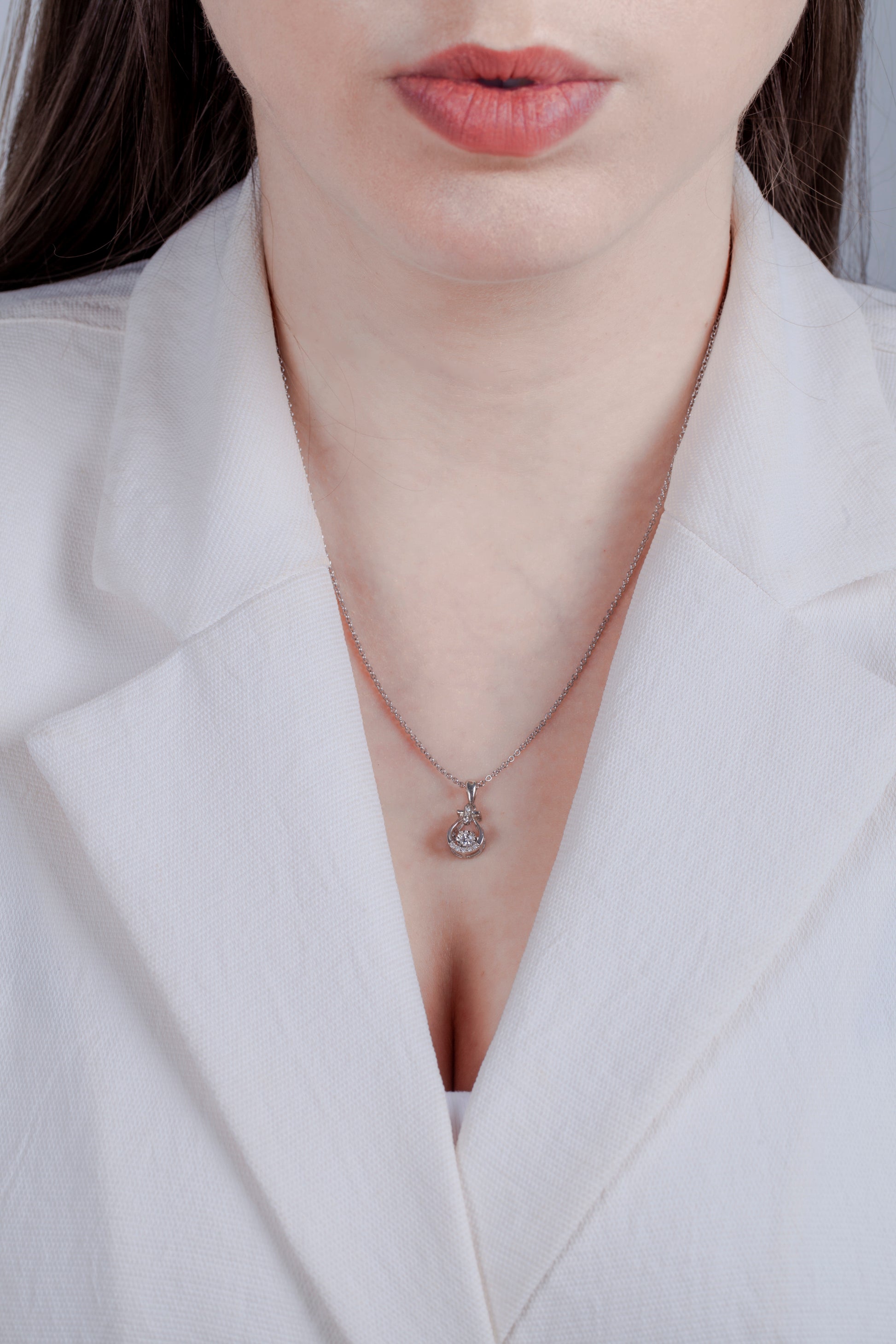 Model in white blazer wearing sterling silver pendant featuring a contoured bell-shaped frame with a structured crossbar at the top. The center holds a dancing solitaire diamond in a miracle setting, accented by three vertical diamonds on the bale and a curved row of diamonds along the bottom edge.