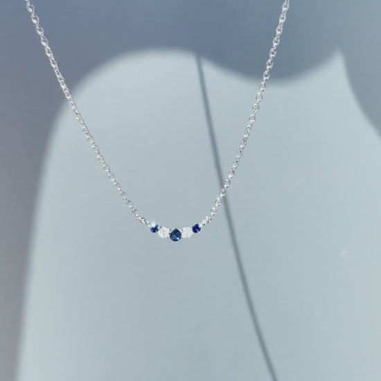 Product showcase video for the .925 Sterling Silver 1/20 Cttw White Diamond & Blue Sapphire Alternating Stone V Shaped 3MM Curve Pendant Necklace. Features close-up details of the alternating sapphire and diamond arrangement (H-I color, SI2-I1 clarity) along the 3mm curved bar, alongside an on-model view demonstrating placement on the 16"-18" adjustable chain.