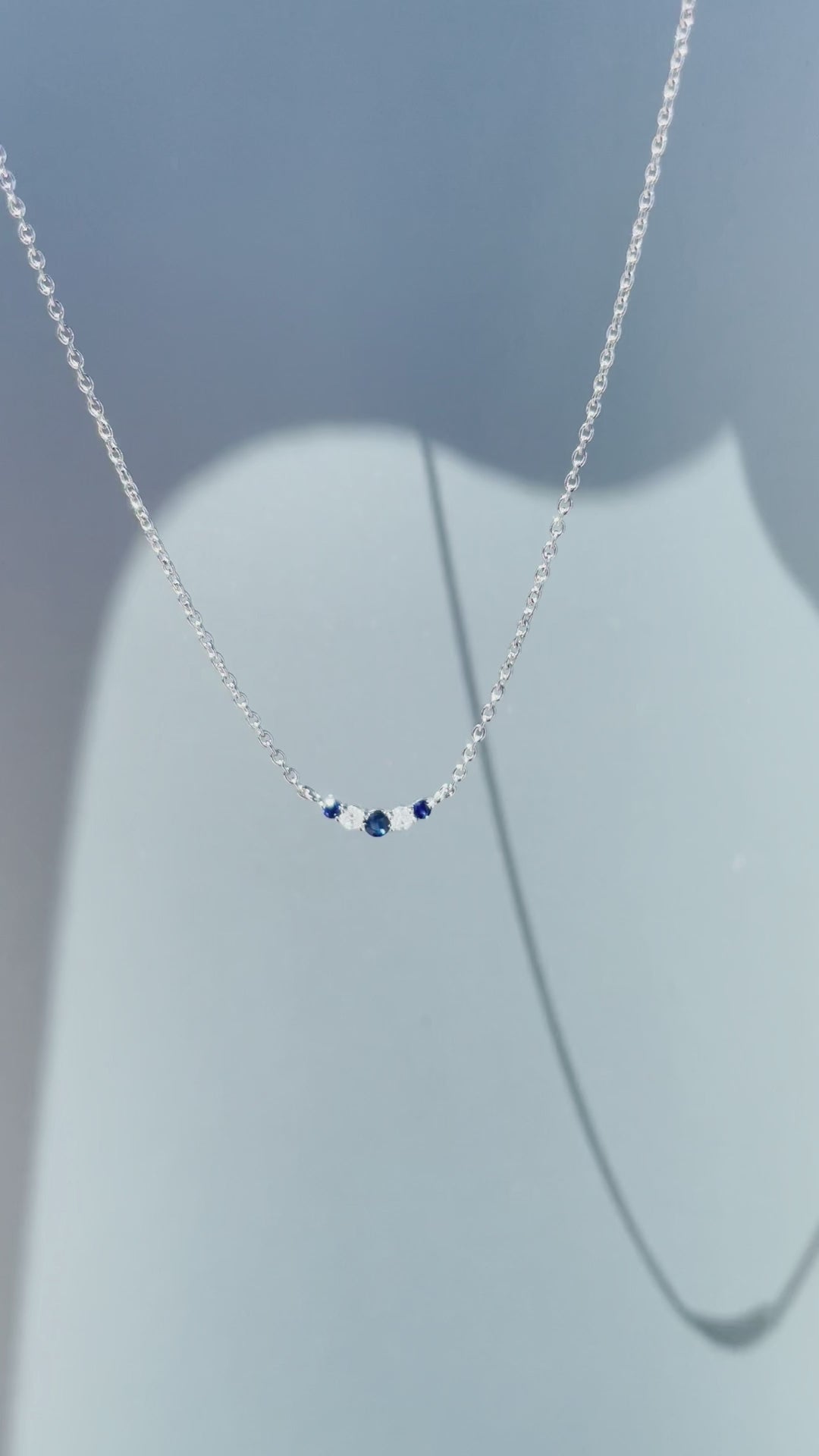 Product showcase video for the .925 Sterling Silver 1/20 Cttw White Diamond & Blue Sapphire Alternating Stone V Shaped 3MM Curve Pendant Necklace. Features close-up details of the alternating sapphire and diamond arrangement (H-I color, SI2-I1 clarity) along the 3mm curved bar, alongside an on-model view demonstrating placement on the 16"-18" adjustable chain.