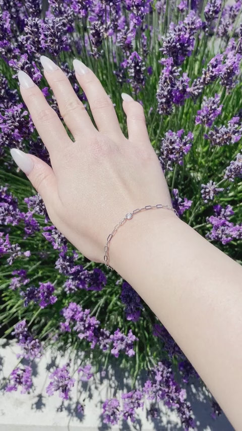 Close-up video of a model's wrist wearing a minimalist silver paperclip chain bracelet. The arm moves gently in bright daylight, catching the flash and brilliant sparkle of a single, central bezel-set Canadian diamond against a rich backdrop of purple lavender.