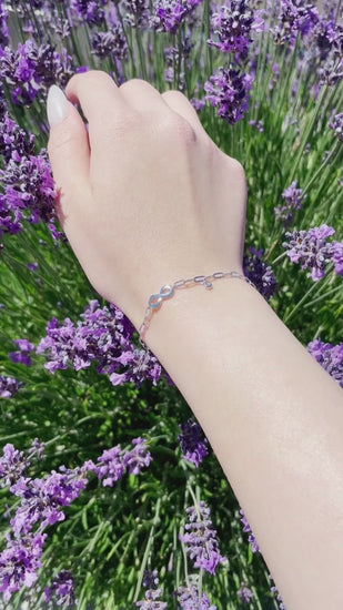 A close-up video of a model's wrist wearing a silver paperclip chain infinity bracelet with a bezel-set diamond charm. The wrist gently rotates in natural sunlight to showcase the metallic shimmer and diamond sparkle against a vibrant background of purple lavender.