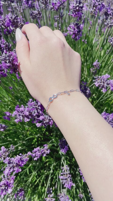A close-up video of a model's wrist wearing a silver paperclip chain infinity bracelet with a bezel-set diamond charm. The wrist gently rotates in natural sunlight to showcase the metallic shimmer and diamond sparkle against a vibrant background of purple lavender.