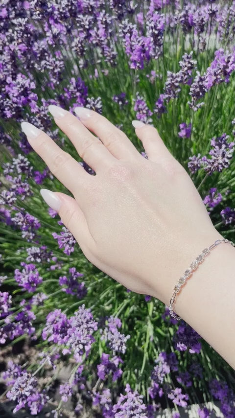 Close-up video of a model's wrist moving in natural sunlight to showcase a silver paperclip chain bracelet. The video highlights the brilliant sparkle of 6 warm Canadian coffee diamonds alternating with 5 white diamonds, set against a vibrant backdrop of purple lavender.