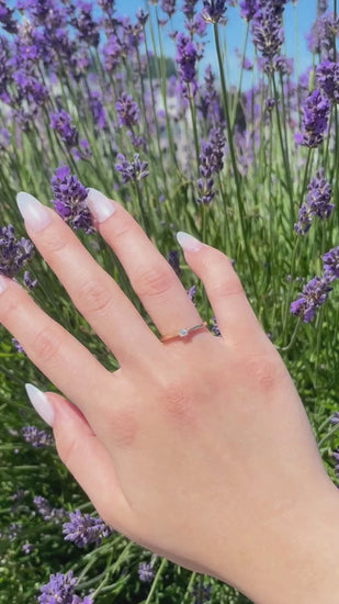 Video showcasing a minimalist 10K yellow gold solitaire ring on a model's hand moving gently in bright daylight. The video captures the sharp flash and brilliance of a single 0.10 ct round brilliant Canadian diamond against a rich backdrop of purple lavender.